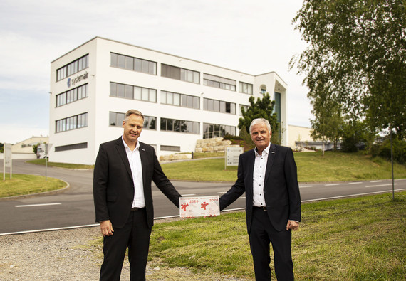Zwei Männer in Anzügen stehen vor einem modernen Bürogebäude und halten gemeinsam ein Schild mit einem roten Kreuz.