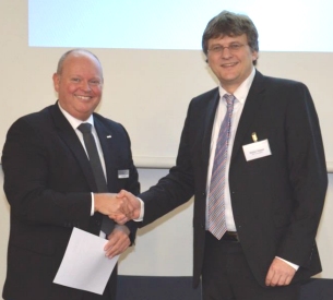 Zwei Männer in Anzügen schütteln sich die Hände vor einem Bildschirm.