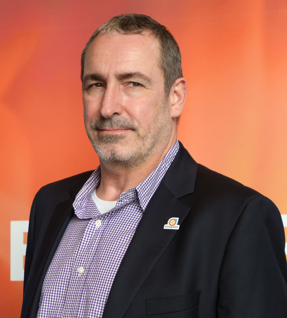 Ein Mann mit kurzem Haar und Bart trägt ein Hemd und einen Anzug, steht vor einem orangefarbenen Hintergrund.