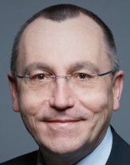 Ein Mann mit Brille und grauem Haar, formell gekleidet, blickt ernst in die Kamera.