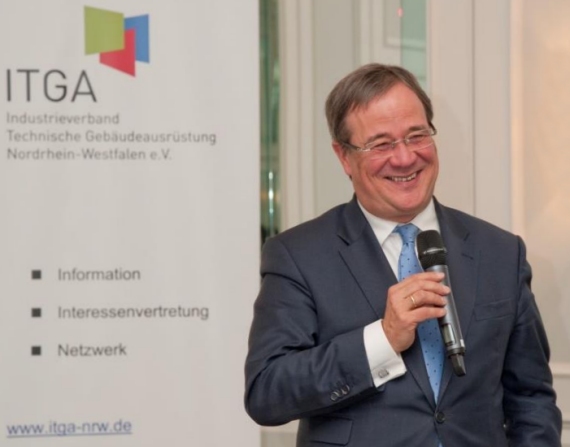 Ein Mann im Anzug hält ein Mikrofon und spricht vor einem Hintergrund mit dem Logo und den Schriftzügen 