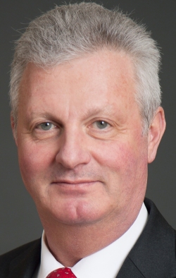 Ein Mann mit grauem Haar und einem ernsten Gesichtsausdruck trägt einen dunklen Anzug und ein weißes Hemd.