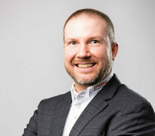 Ein Mann mit kurzen blonden Haaren und Bart trägt ein weißes Hemd und einen dunklen Anzug, lächelt und steht vor einem unscharfen Hintergrund.