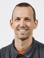 Ein Mann mit kurzen Haaren und Bart trägt ein graues Hemd mit orangefarbener Krawatte und lächelt.